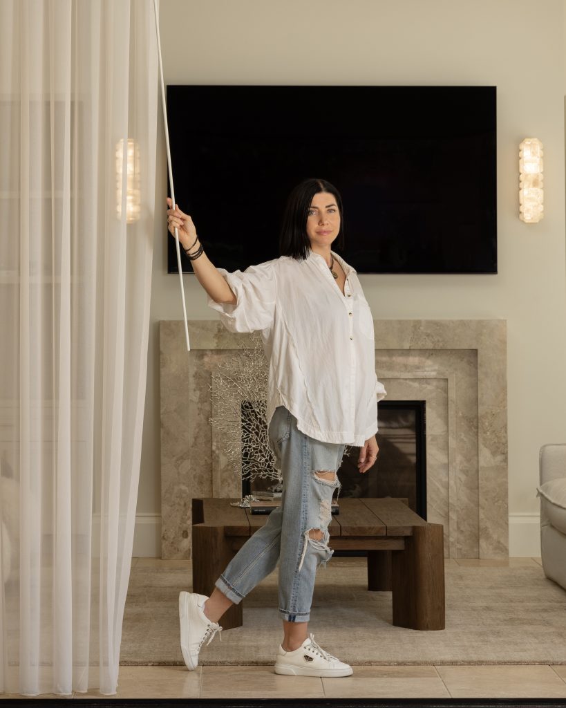 Woman in a white shirt and ripped jeans stands in a living room, holding a curtain rod beside a fireplace and large TV.